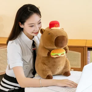 A medium-sized capybara plush toy wearing a red hat and holding a plush hamburger, sitting on a desk next to a smiling person.