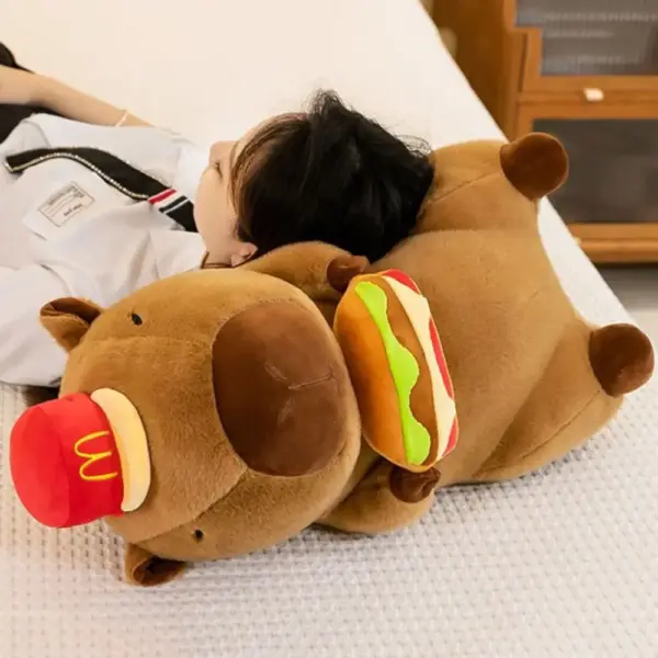 A large capybara plush toy wearing a red hat and holding a plush hamburger, being used as a pillow by a person lying down.
